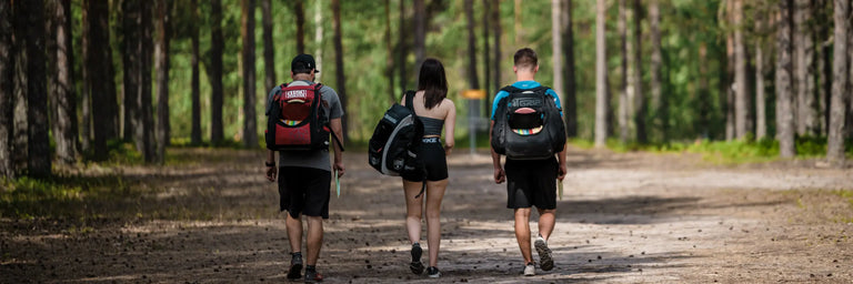 tre disc golfere går med tasker på en disc golf bane i skoven