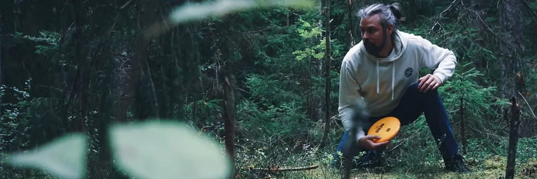 Kastaplast sponsoreret disc golfer der sidder på hug i skoven med en hvid hættetrøje gør klar til at putte